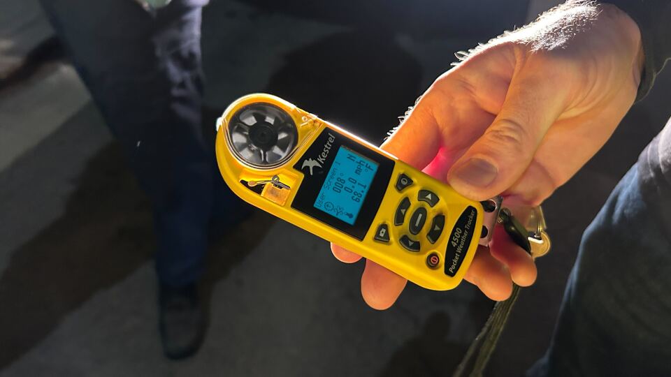 A close up of a man's white hand holding a yellow device with fan blades on it that can measure wind speed. The device's blue screen shows direction, speed and temperature.
