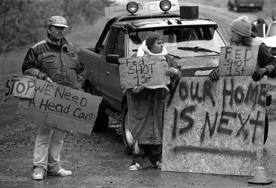 A black and white photo of people holding up cardboard signs that read "Stop. We need a head count," "Fed shot 1st," and a sign on plywood that reads "Your home is next." They stand in front of a truck on a dirt road.