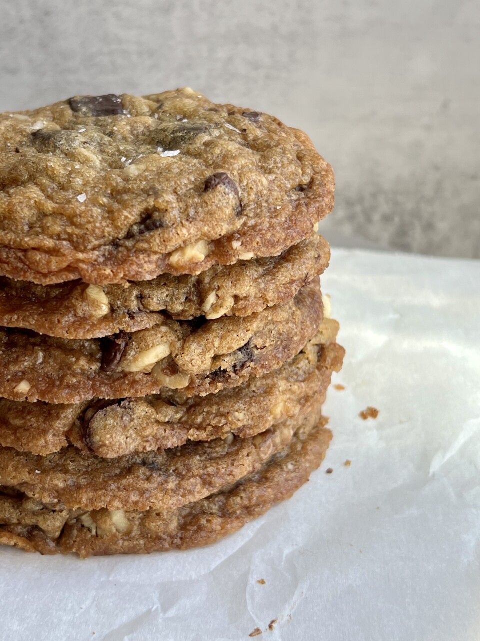 A photo of six walnut chocolate chip cookies are perfectly stacked one on top of the other on a white plate, with a few delicious crumbs off to the side. The cookies are finished off with bright white flakes of sea salt.   