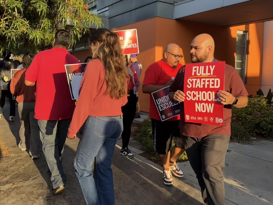 A group of adults walk in a small circle outside a building. They all wear red shirts and carry signs. One visible sign says "Fully staffed schools now" in all capital letters.