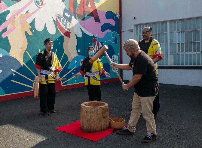 A man with medium dark skin tone in casual clothing raises a wooden mallet to pound mochi while three Kodama Taiko performers in traditional happi coats look on, with a large wooden mortar placed on a red mat in front of a vibrant mural.