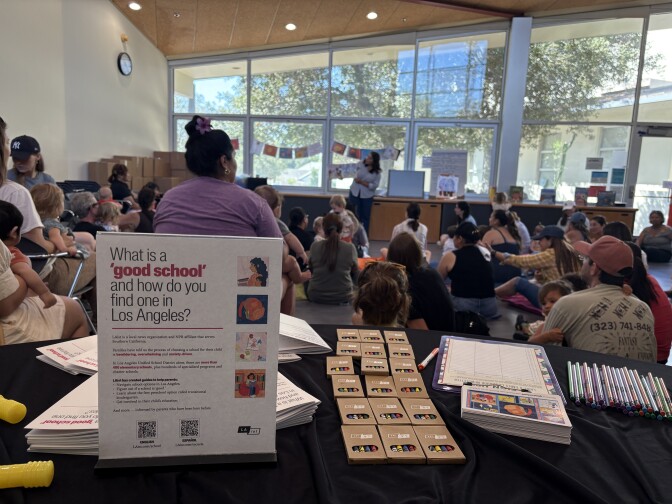 Colorful crayons, post cards, pencils and workbooks are displayed on a table with a black table cloth. The backdrop is parents and caregivers sitting on the floor, and some in chairs, with their toddlers and babies listening to a librarian read a children's book. 