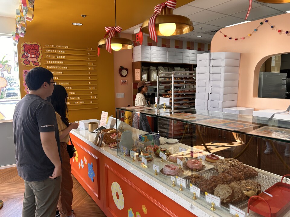 Two people, a man and a woman, are standing in front of an orange counter filled with doughnuts, protected by a glass case. A woman wearing a white shirt and brown hat can be seen in front of them, walking back towards the kitchen, which isn't pictured.