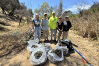 A group of five people stand side by side on a dirt path with seven large plastic bags full of plant material. 