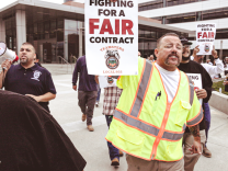 A man in a neon vest is walking and holding a sign that reads "For a Fair Contract Teamsters Local 952". Crowds of people behind him are also walking and holding signs.