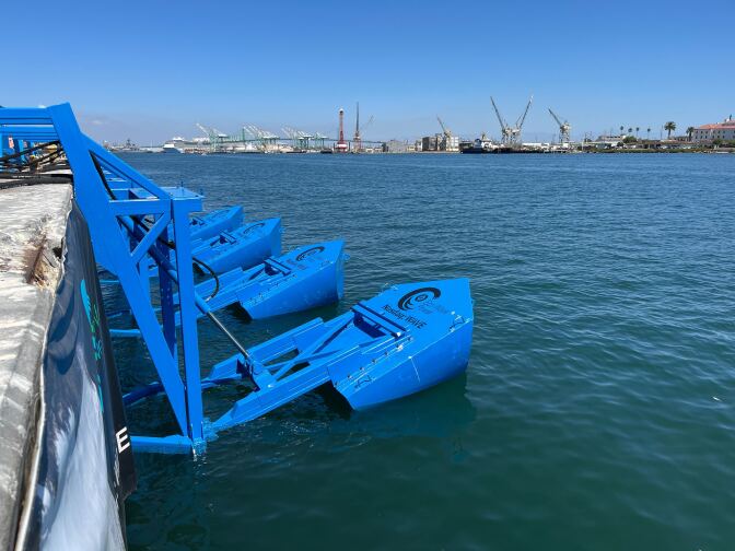 Blue machinery sits on the water's surface, with port infrastructure across the water beyond.