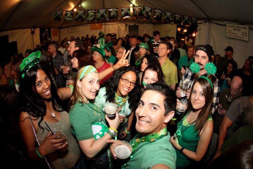 Patrons of the Tam O'Shanter pub take a cheerful group selfie of their St. Patty's Day celebrations in their green outfits while holding cups of Guinness beer.