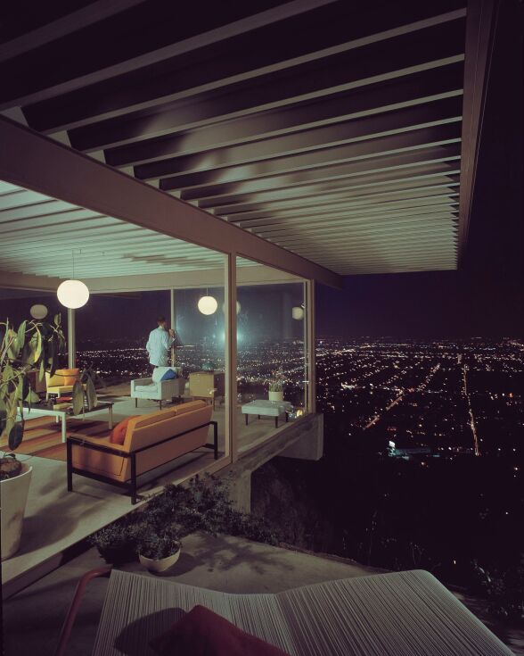 A color photo of a modernist house at night. A man in a suit jack is standing at one of the large windows, looking at the night view below.
