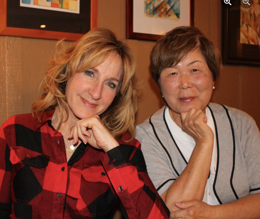 A long-haired white blonde woman in a red and black plaid shirt poses seated next to an Asian American woman who wears her brown hair short and a short-sleeved gray sweater with black piping.