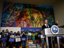 Gov. Newsom in a suit and tie stands at a lectern in front of a group carrying signs supporting him.