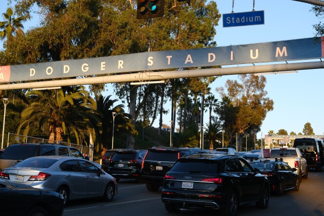 Cars are backed up across four lanes heading into Dodger Stadium, under a Dodger Stadium banner that notes it's at GATE A, with a street sign reading STADIUM above, and traffic lights to the left of that.