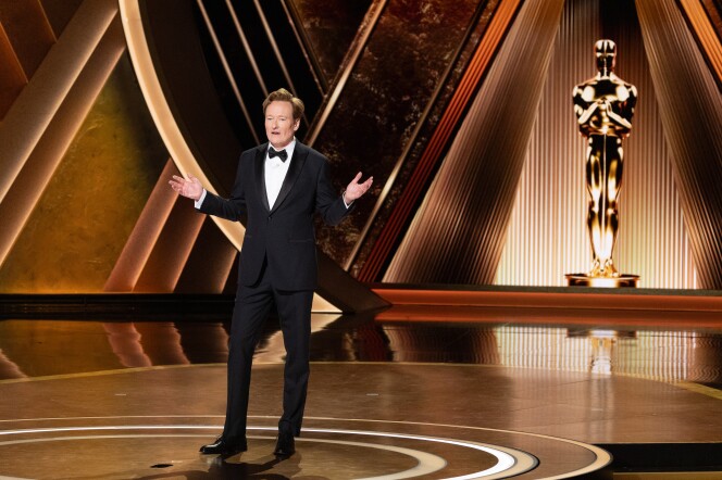 Conan O'Brien stands on the Oscars stage wearing a black tux with a bow tie. His arms are outstretched at the elbows as he speaks to the audience. A tall golden Oscar statue is behind him on stage.