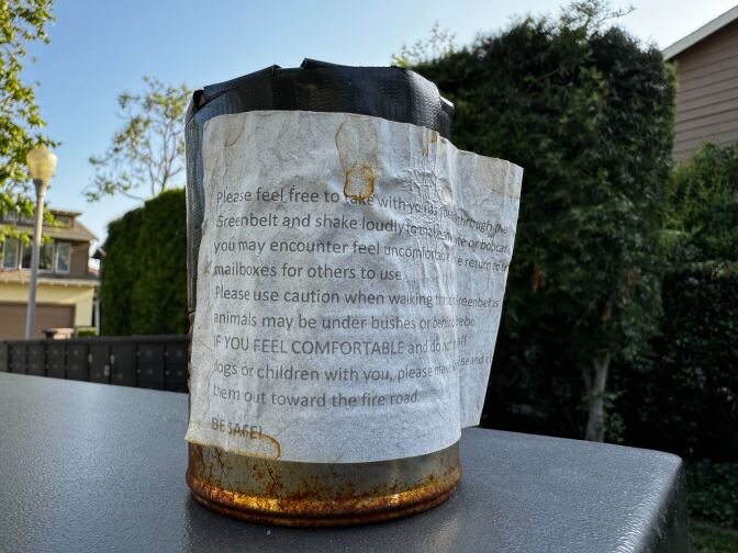 An old rusty can sits on the corner of a table, surrounded by large hedges. On it there's an old stained piece of paper with text explaining what the can is used for