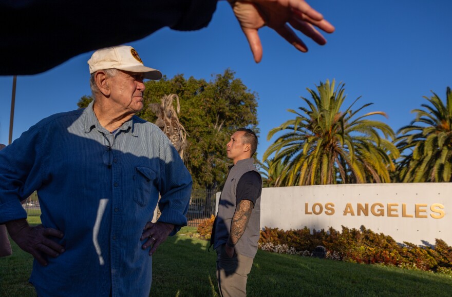 An older man with light-tone skin wears a ball cap as he looks to the left. A person's hand is gesturing at the top of the frame. Palme trees are in the background and a sign reads: Los Angeles