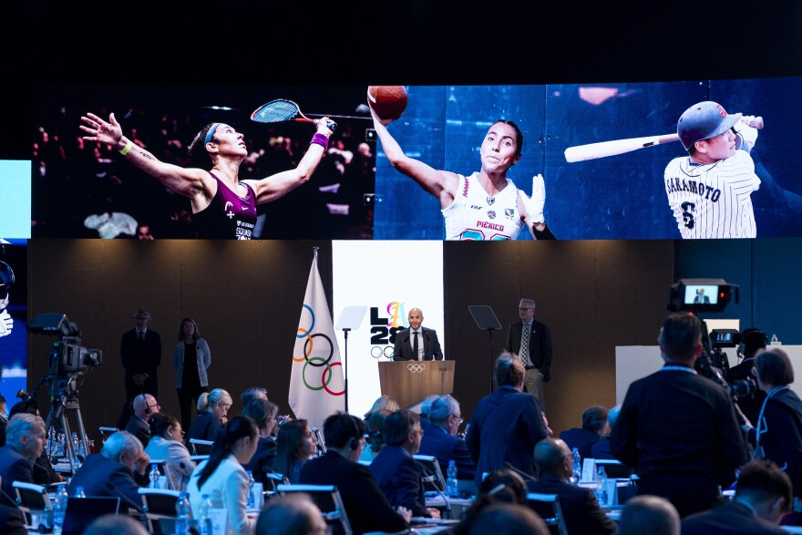 A screen shows three images of athletes in varying stances of playing a sport. A man stands at a podium with the Olympic flag behind him. People who are seated look at the screens and at the podium.
