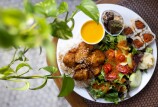 A white plate sitting on a table top that contains chicken over a bed of rice, a small cup of yellow soup, a paper cup of cookies, and green salad with small sliced tomatoes covered in dressing. To the left is a green plant vine.