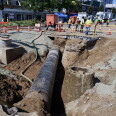 A section of a city street has been dug up, creating a deep trench with an exposed pipe running down the middle. 