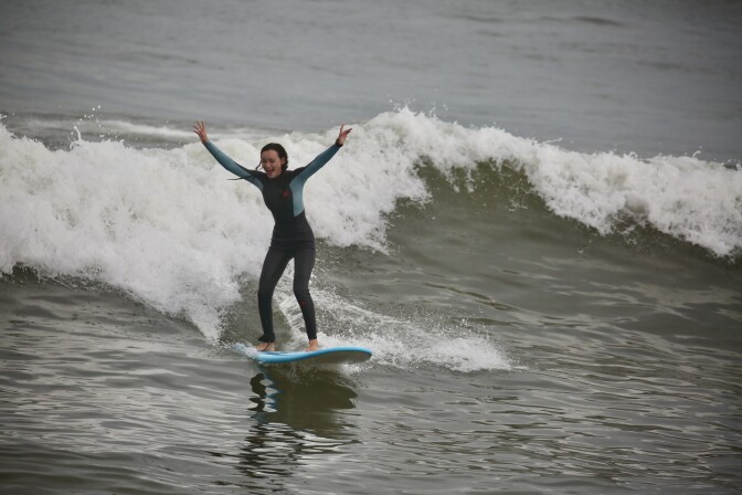 A young woman smiled wide and holds her hand in the air with surfing a wave. She has long hair and is wearing a black wetsuit with blue on the sides. 