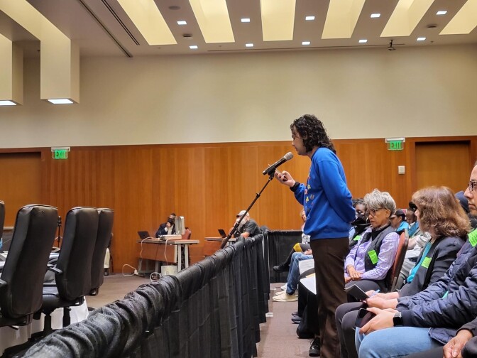 A man with brown skin and wearing a bright blue sweatshirt stands at a microphone in an auditorium filled with people.
