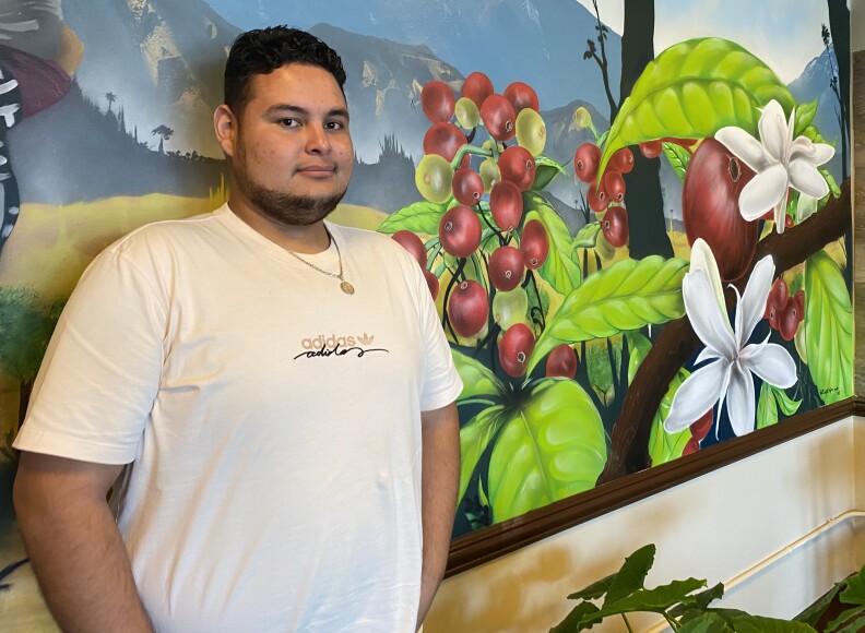 Irving Alvarez, 28, stands in front of a mural of a mountainscape with colorful white and red flora in the foreground. Irving wears a white Addidas t-shirt and a gold medallion chain.