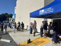 People wait in line under the sun beside an off white building. There's a shade ten with blue awning and white lettering "LADWP"