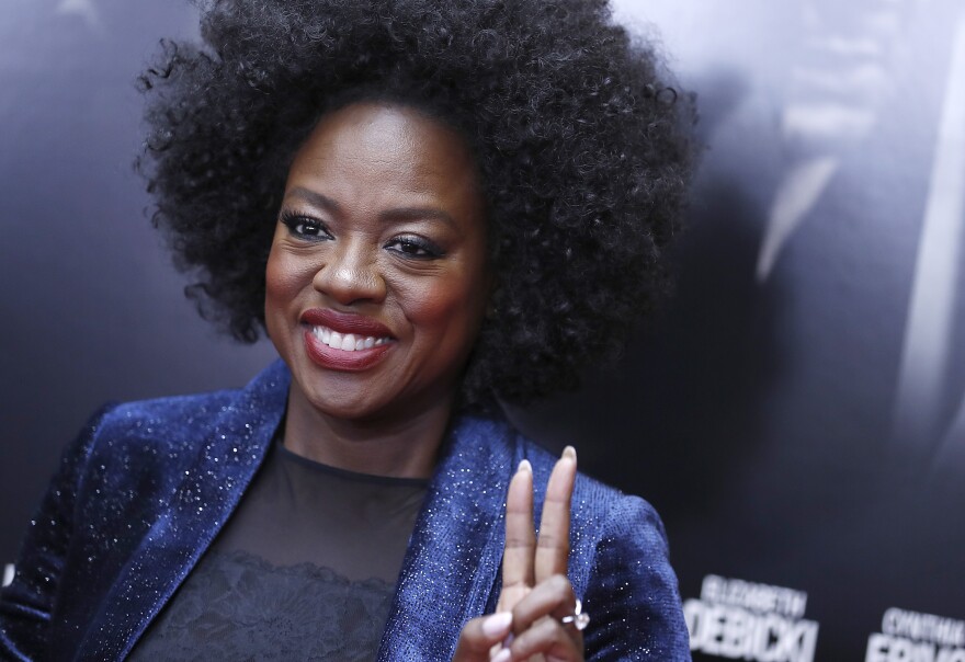 Actress Viola Davis smiles and flashes a peace sign on a red carpet. She is dressed in a sparkling navy blazer and sheer navy top.