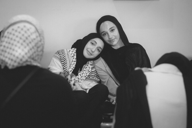 A black and white photo of two teen girls wearing black hijabs sitting against a wall. One girl, to the left of frame rests her head on the shoulders of the other. She also wears a Palestinian keffiyeh around her shoulders.