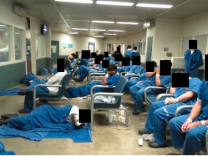 A room with white and green walls and bright lights is crowded with men whose faces are obscured in blue jail uniforms. Some are sleeping on the floor wrapped in blue blankets. Others are sleeping upright in chairs. 