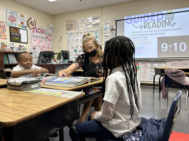A teacher with light-toned skin sits pointing to a book. Two young Black girls sit on either side of the set of desks. A projector in the background has a slide that says "GUIDED READING; 9-10" with other details in smaller print. The walls are decorated with various learning aids.