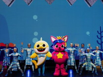 dancers surround two performers in baby shark and cat costumes on a stage.