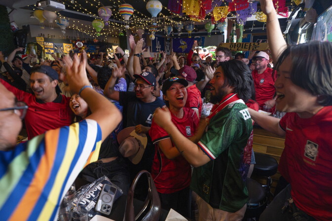 Large group of people standing and cheering inside of a restaurant. People are dressed in different team soccer jerseys.