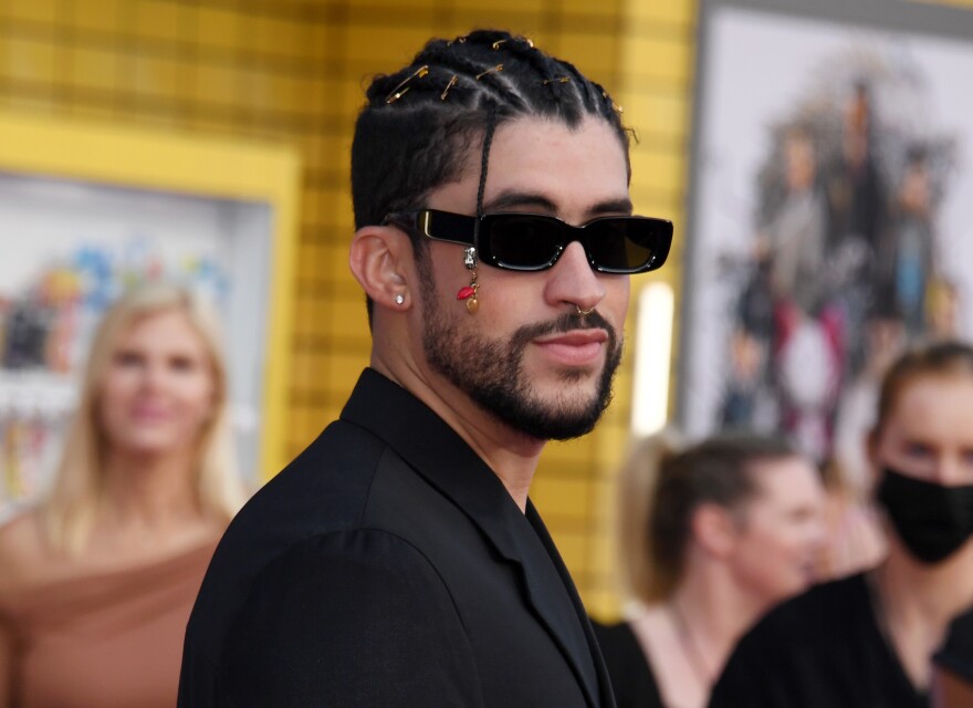 Bad Bunny is dressed in a black suit and wearing dark sunglasses. Other people appear in the background, and one woman is wearing a black facemask.