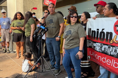 A man with medium skin tone speaks into a microphone. In front of him, there is a stand with several other microphones, each with a mic flag representing a  news organizations. About a dozen people stand around the man in the center, many wearing forest green shirts that read "UNION DEL BARRIO." On the ground, there is a small child playing with toy cars. 