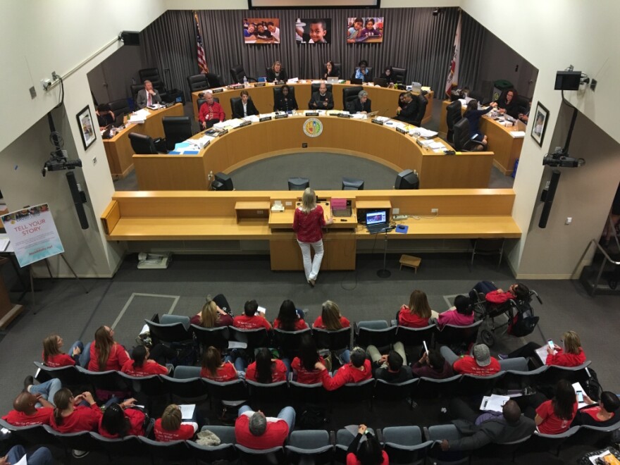 An overhead shot of a space where a public meeting — a meeting of the L.A. Unified School Board — is taking place. There's a half-circle-shaped dais at the front of the room where several public officials are seated. The front rows of theater-style seats below are filled with people wearing red. One of the red-clad attendees of the meeting is addressing the board.