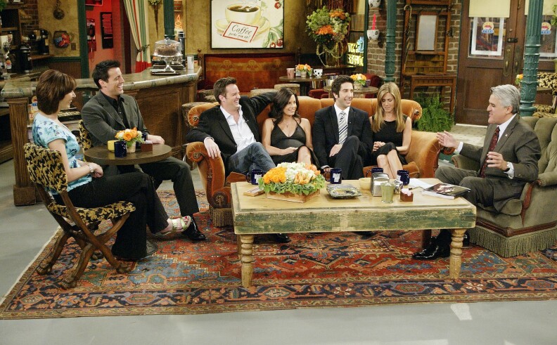 Seven people are seated on TV show set that resembles a living rom with a patterned rug, a heavy table, and a sofa and chairs arrayed around the table.