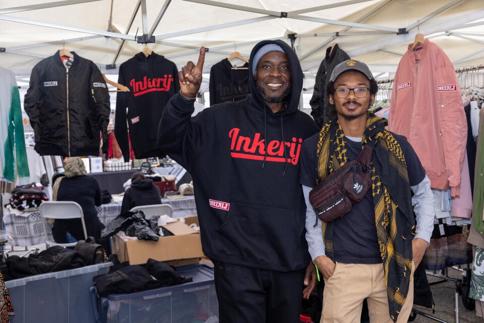 A Black man wearing a black hoodie with the words Inkerij in red holds up his hands in the peace sign beside another Black man wearing a black t-shirt and keffiyeh. 