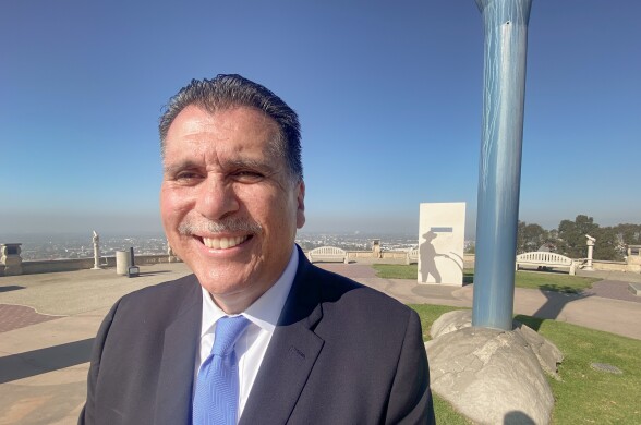 A man in a dark suit and blue tie smiles for the camera. Behind him we see a park and some sort of public monument.