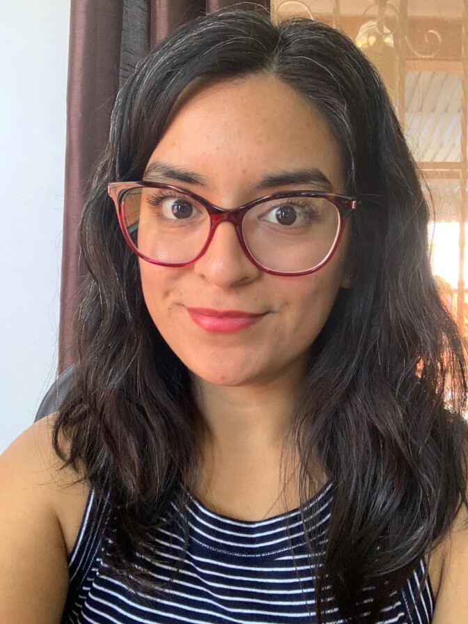 An image of a woman's face. She wears red-rimmed glasses, a striped black-and-white sleeveless shirt, and has dark hair. 