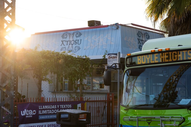 The exterior of a restaurant painted baby blue with the lettering that reads "X'tiosu." Next to the store front on the street, to the right of frame a green bus passes by with a sign that reads "Boyle Heights."