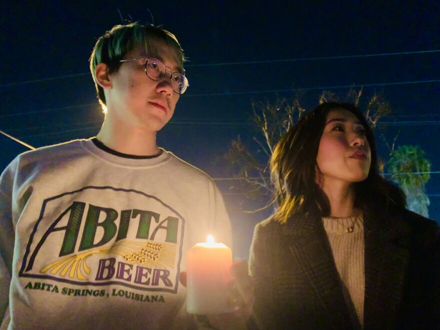 A Chinese American man wearing glasses and a sweatshirt that reads "Abita Beer" holds a candle, while standing next to a Korean American woman wearing a pea coat over a sweater.