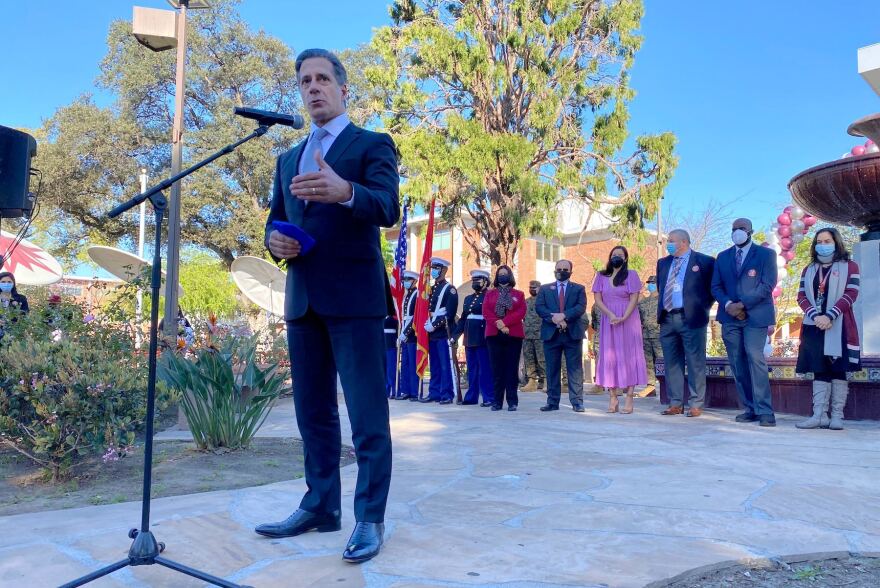 A man in a blue suit stands outside at a microphone on a clear day. A row of official looking people stands lined up behind him. A tree and a fountain are behind them.