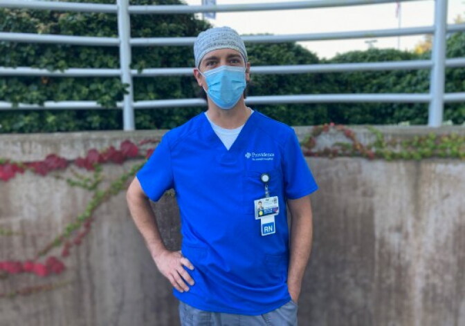 A person in bright blue scrubs and blue mask stands with hand on hip outside. Their is a ID with an RN designation hanging from the shirt pocket.