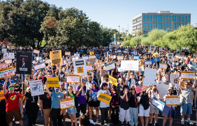 A crowd of people gathers, holding yellow signs that read "Stand With Animation!" and handmade signs. The ground stretches into the background, where trees that frame the street. One building is visible in the back right corner of the frame.