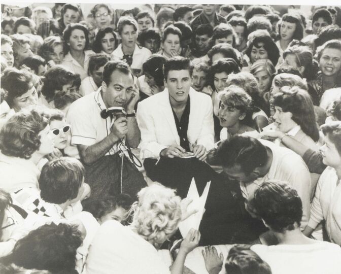 In a black and white photograph, two men sit in in the middle of a throng of young women. The women are all turned towards the men. One of the men wears a white short-sleeved shirt and holds a microphone. The other wears a white sports jacket over a black shirt. 