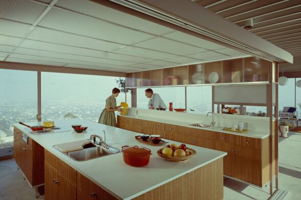 A color photograph of a couple in the kitchen of a sleek mid-century modern house.