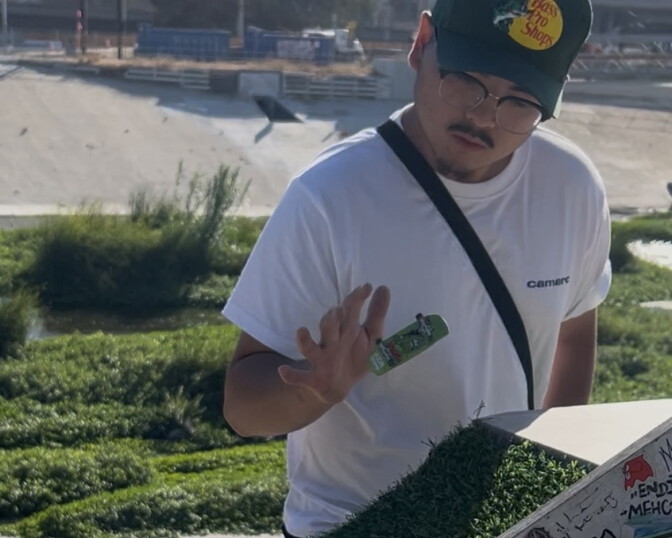 Jimmy Tran wears a green "Bass Pro Shops" cap and a white t-shirt as he performs a trick in mid-air at the LA River Fingerboard skatepark. The bottom of the LA River and the other side of the embankment is seen behind him.