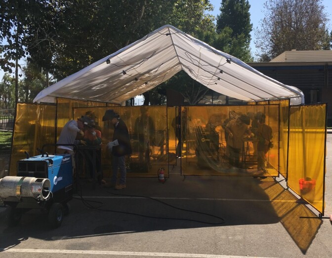 A white tent in a parking lot provides shade to students who are practicing their welding. 