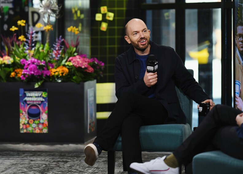 a bald man in black clothes holds a microphone while sitting in a chair