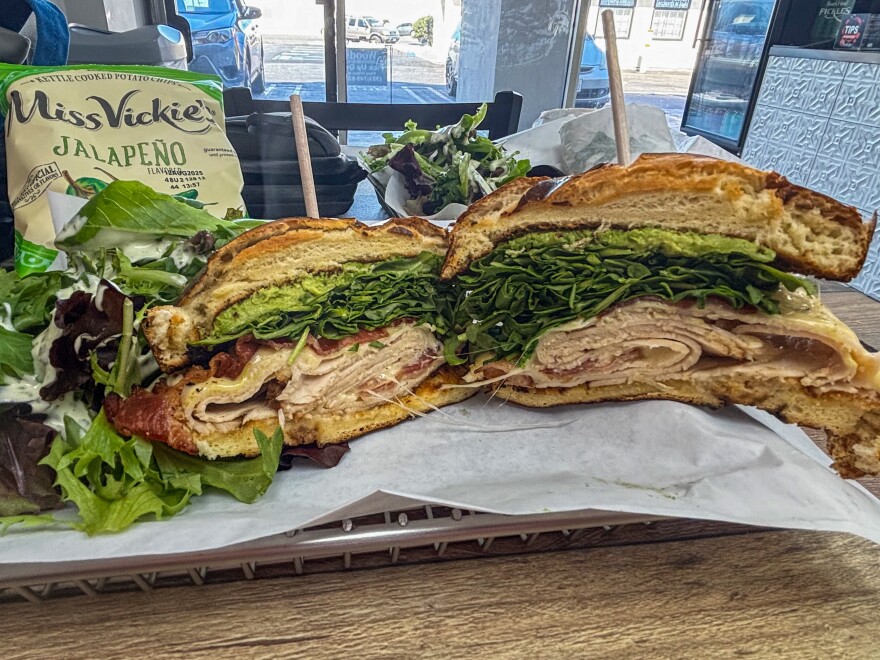 A large sandwich cut in half on a tray, filled with layers of sliced turkey, crispy bacon, leafy greens, melted cheese, and avocado spread on toasted bread. Served with a side salad and a bag of Miss Vickie's Jalapeño kettle-cooked chips.