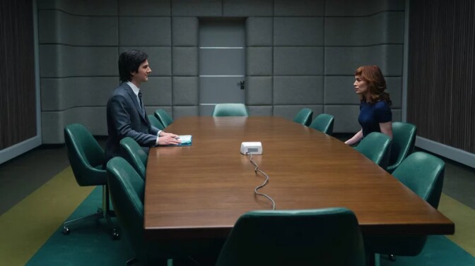 A woman and man who both appear to be white and in their 30s or 40s sit across from each other at an otherwise empty conference table. The room is sterile and empty, and both people wear business attire. 